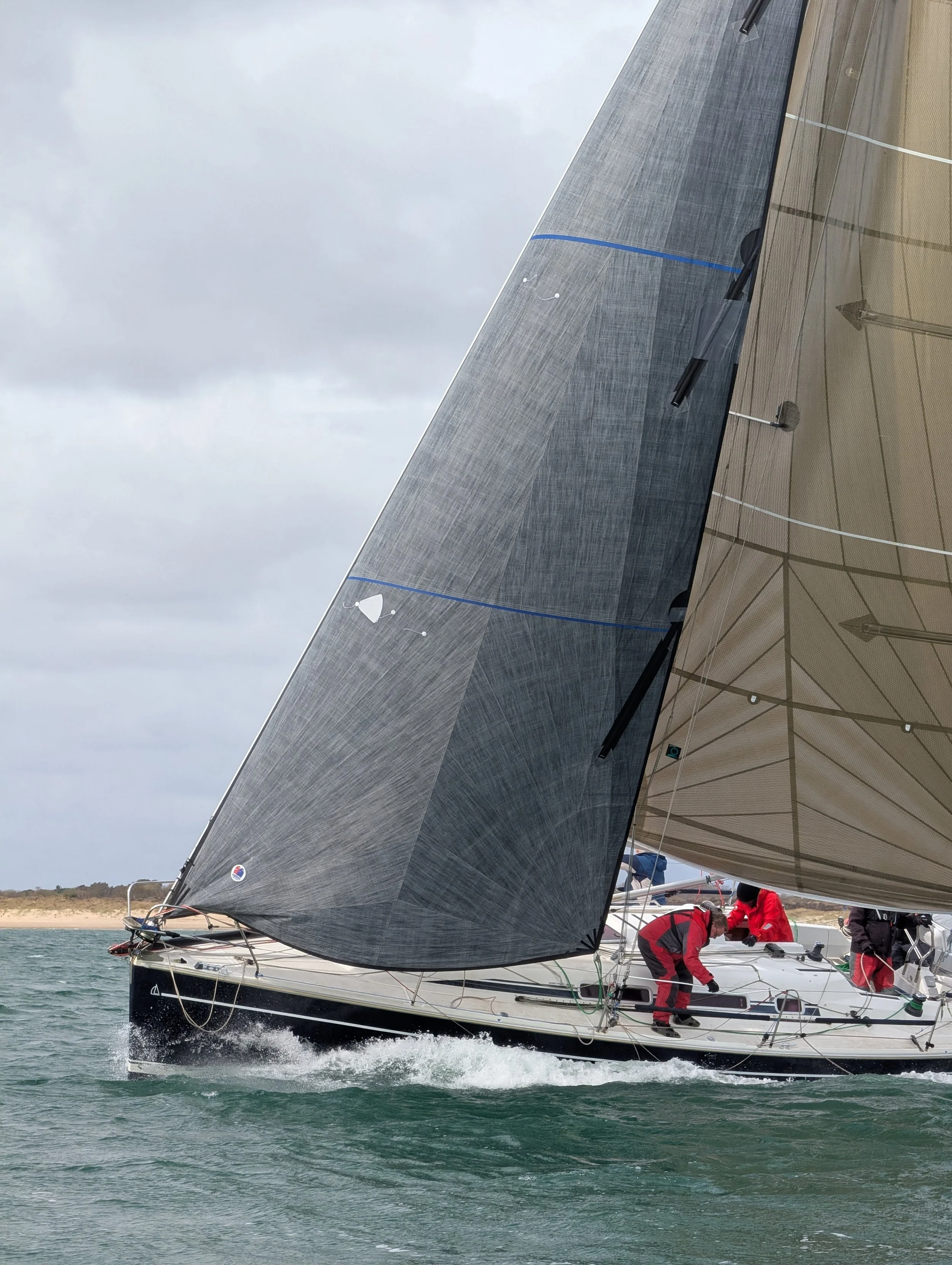 A sailboat with dark sails moving through the water, crew members on deck adjusting equipment, under a cloudy sky.