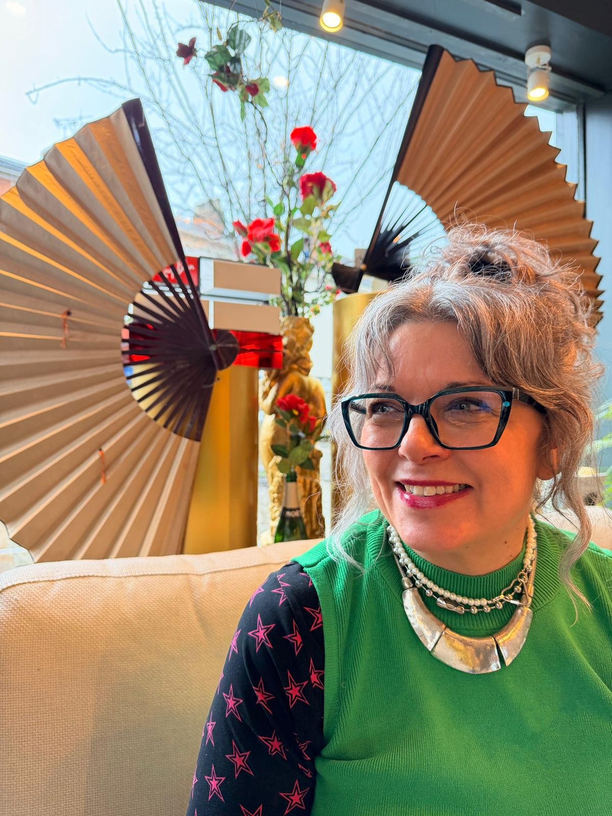 A woman with glasses smiling, wearing a green top, sitting indoors with decorative fans, flowers, and a window behind her.