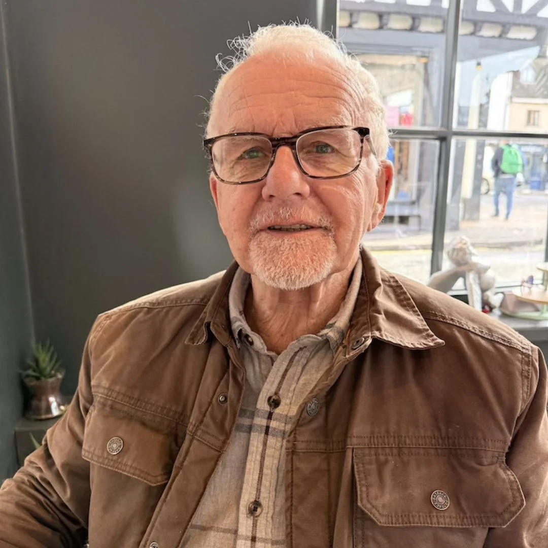 A smiling elderly man with glasses and a white beard, wearing a brown jacket over a plaid shirt, sitting indoors near a window with a city street scene in the background.