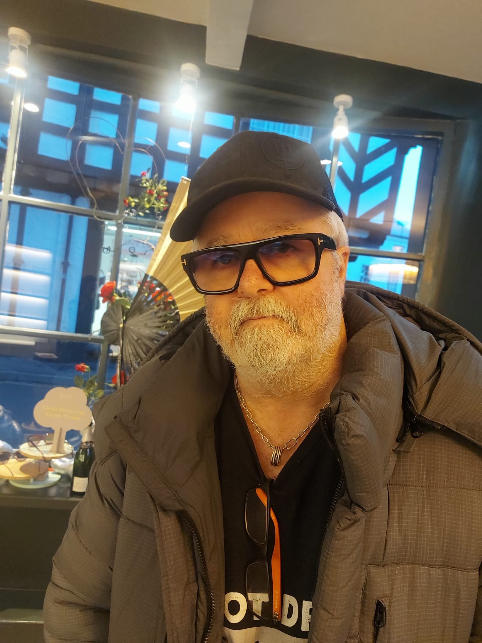 A middle-aged man with a white beard and glasses wearing a black cap, a black hoodie, and a brown jacket. He is indoors with a building or storefront visible behind him, including a display of flowers and a window.