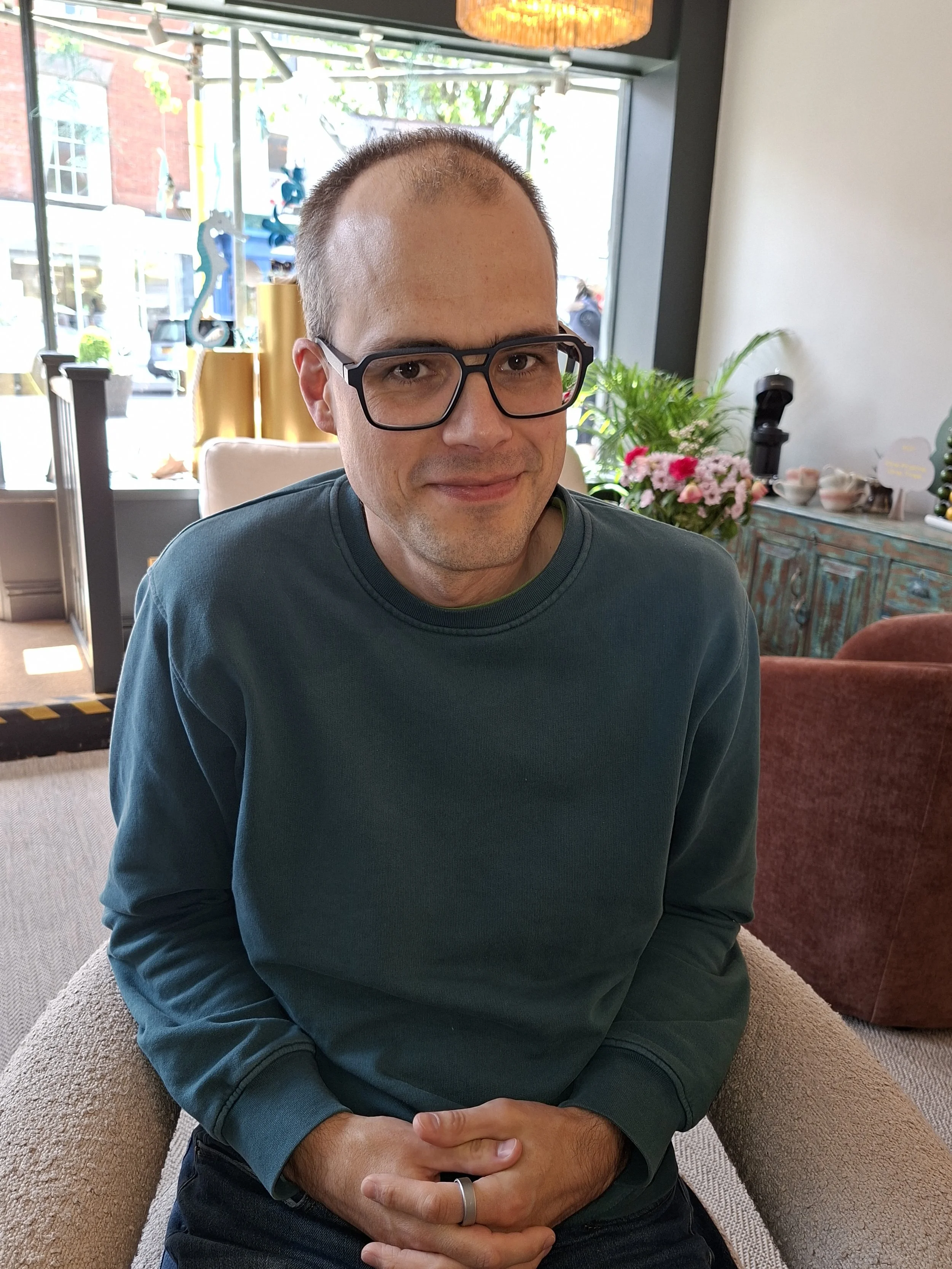 A man with short hair and glasses sitting on a beige armchair in a cozy indoor space, with a background of a window showing a street scene and a wooden sideboard decorated with pink and white flowers, bowls, and plants.
