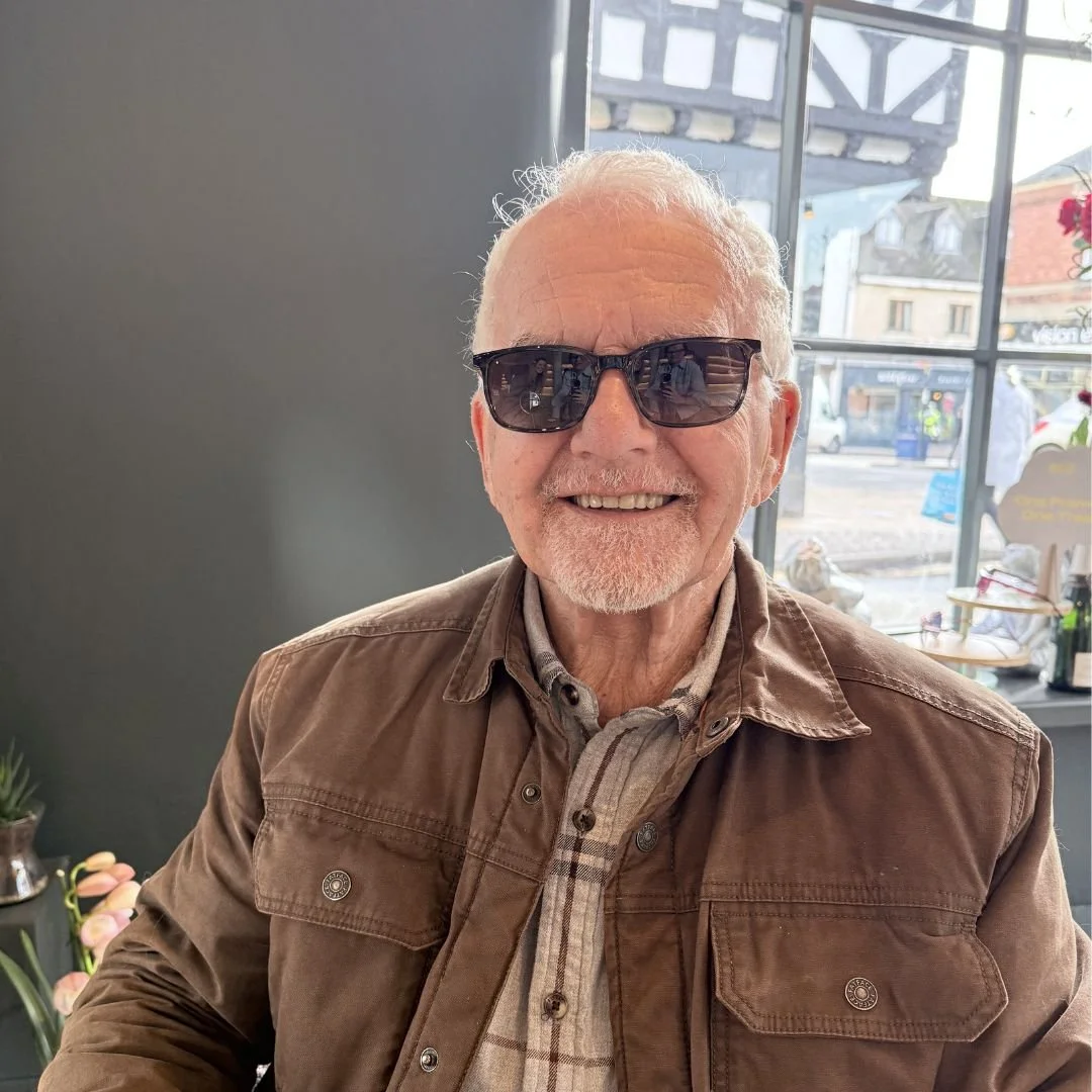 An elderly man with white hair and a beard wearing sunglasses, a beige plaid shirt, and a brown jacket, smiling indoors near a window with a view of a street outside.