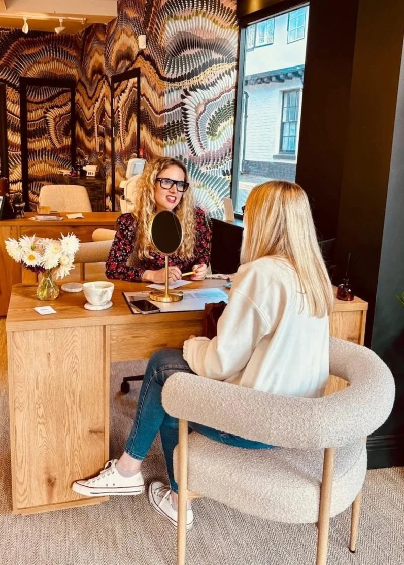 A woman with long blonde hair, wearing a white hoodie and blue jeans, sitting in a beige chair across from a woman with curly blonde hair, glasses, and a floral dress, behind a wooden desk with flowers and a mirror. The room has decorative wallpaper and a large window.