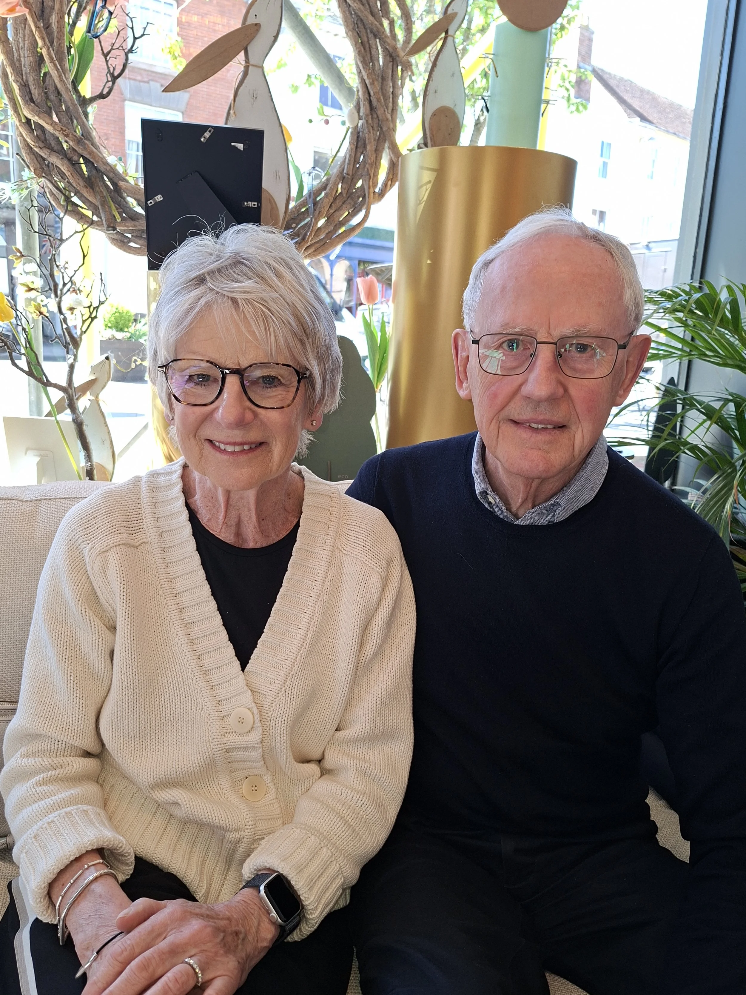 A smiling elderly woman with short gray hair and glasses, wearing a beige cardigan, sitting next to an elderly man with glasses, short gray hair, and a dark sweater, in a well-lit indoor space with plants and decorative elements in the background.
