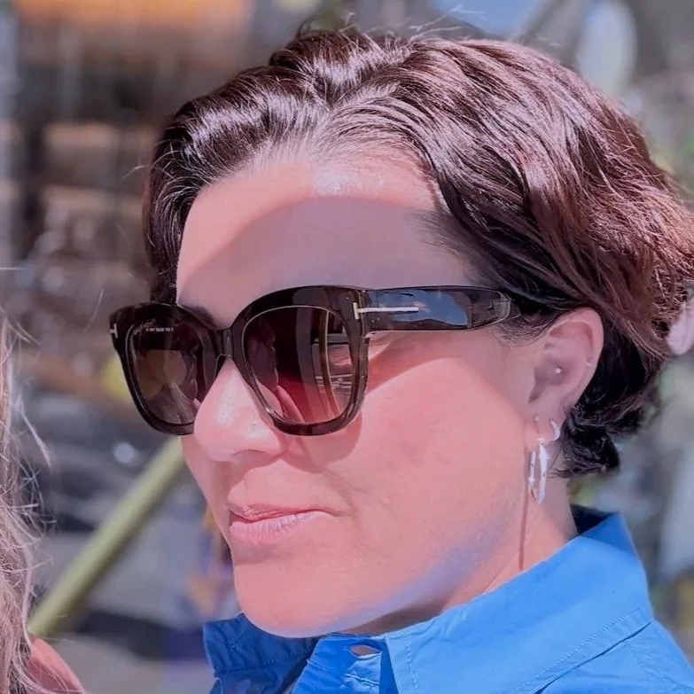 Woman with short brown hair and sunglasses outdoors in bright sunlight, wearing a blue top.