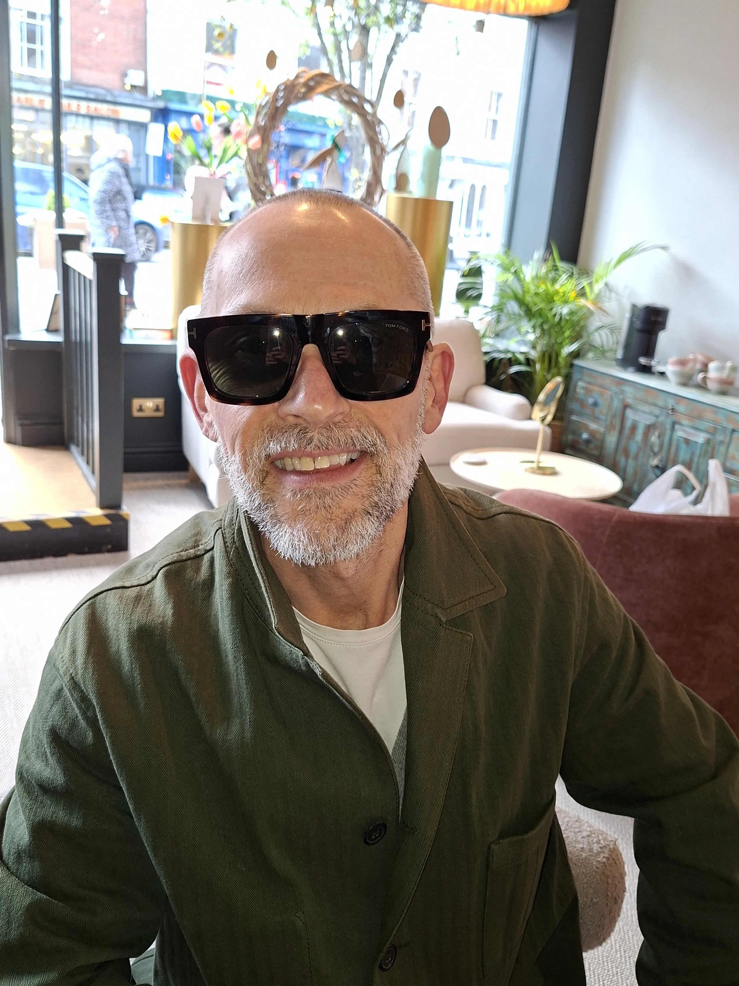 A man with a beard and bald head wearing black sunglasses and a green jacket taking a selfie inside a cafe, with flowers and decorative items visible behind him.