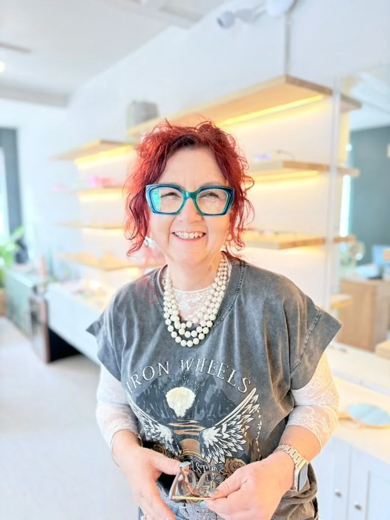 A woman with curly red hair, glasses, and pearl necklace smiling in a bright store with shelves in the background.