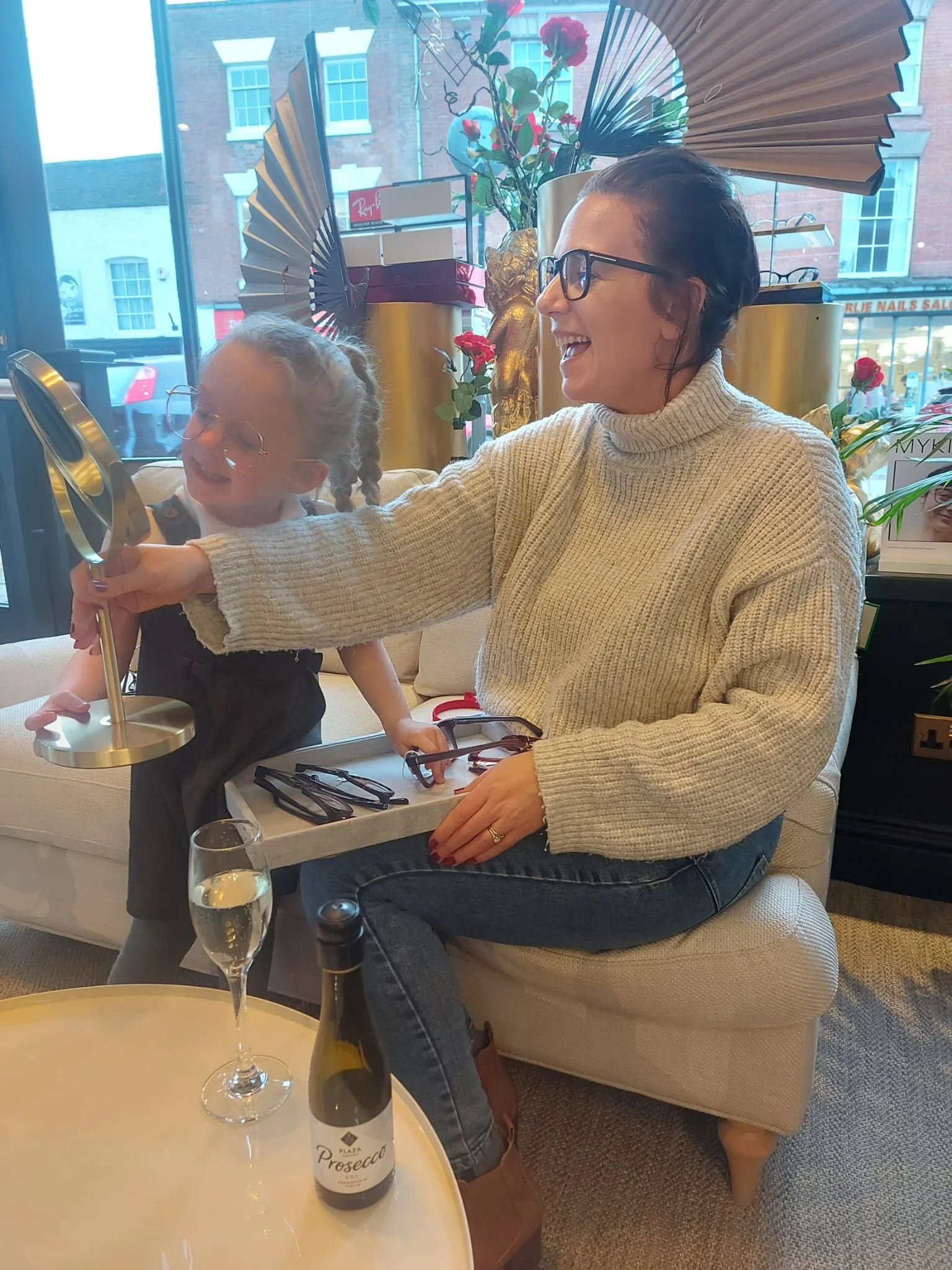 A woman with glasses and a cream sweater smiling and looking at a young girl with glasses and braided hair, who is reaching for a mirror in a brightly decorated room with large windows and various decor items.