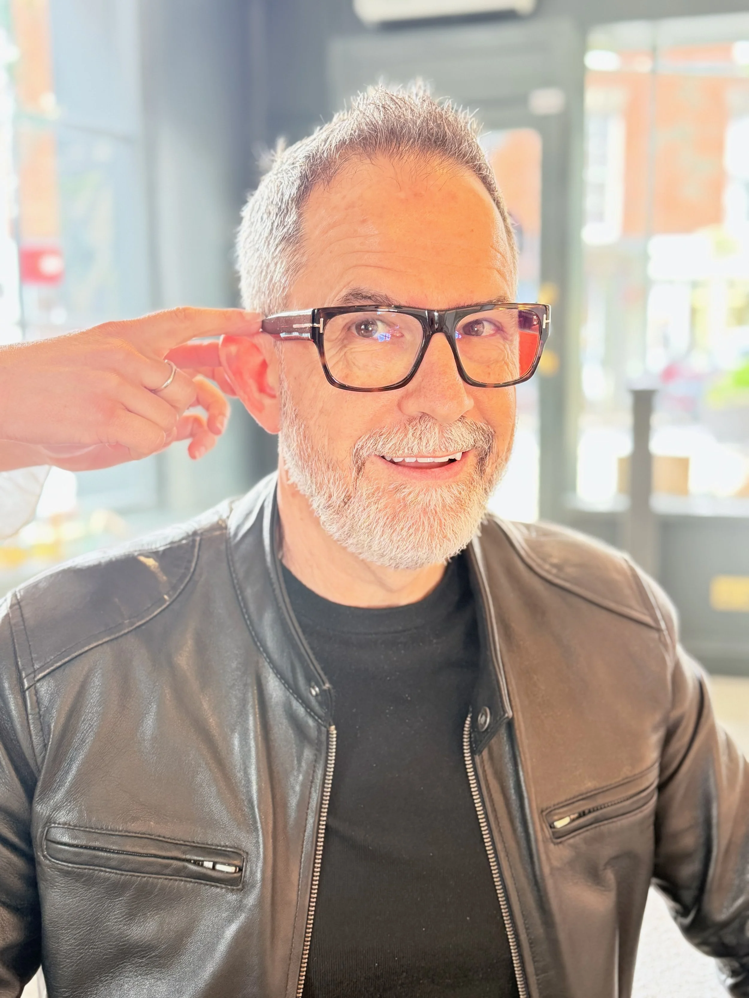 A man with short gray hair and a beard, wearing glasses, a black t-shirt, and a brown leather jacket, adjusting his glasses with his right hand, inside a building with large windows in the background.