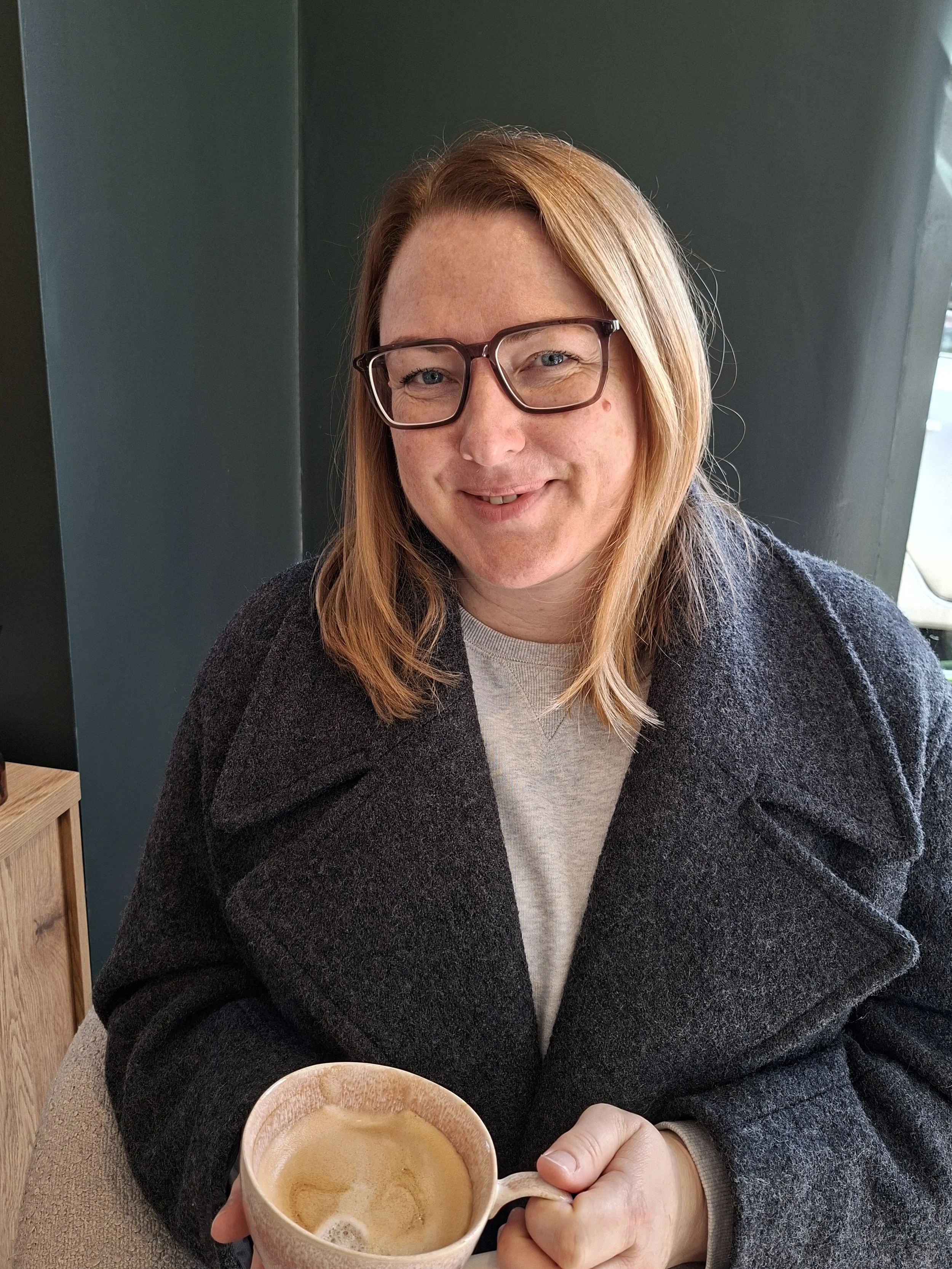 A woman with red hair, glasses, and a warm smile, sitting indoors, holding a mug of coffee. She is wearing a dark coat over a light-colored sweater, with a dark green wall in the background.