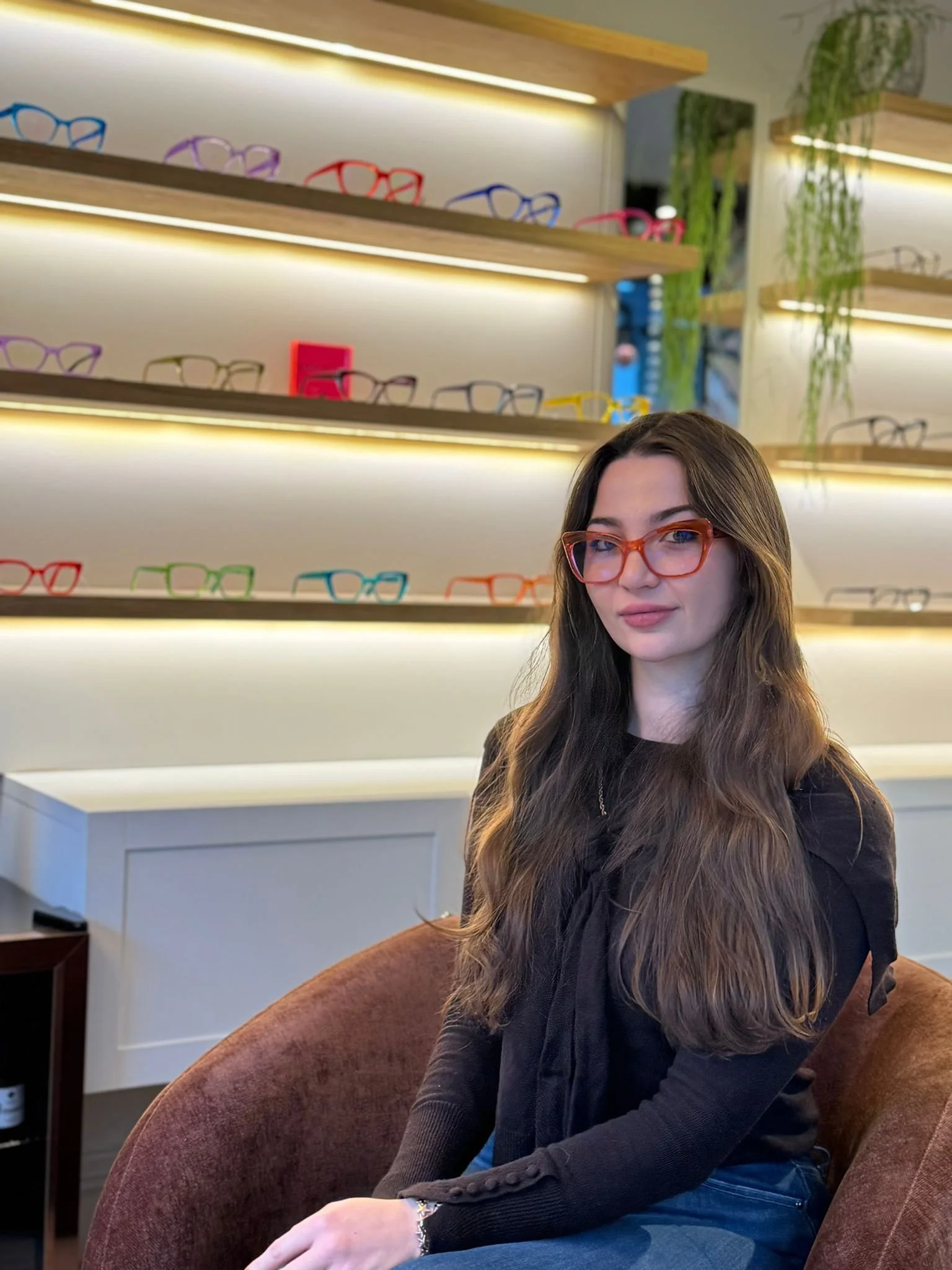 A woman with long brown hair wearing red glasses, a black top, and blue jeans, sitting on a maroon armchair in a retail store, with shelves of colorful eyeglasses and green plants in the background.