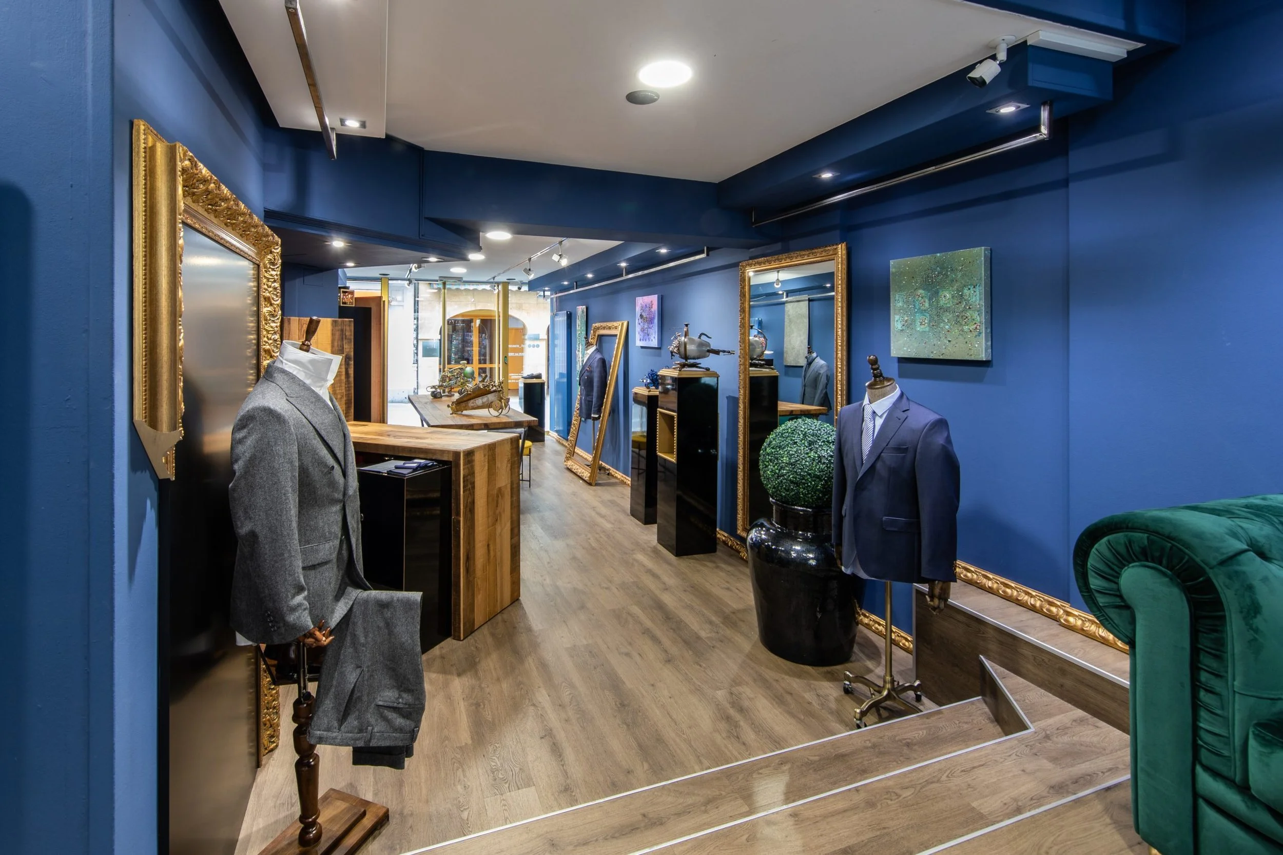 Intérieur d'une boutique de vêtements avec des costumes sur mannequins, des tableaux accrochés aux murs bleus, et un sol en bois.