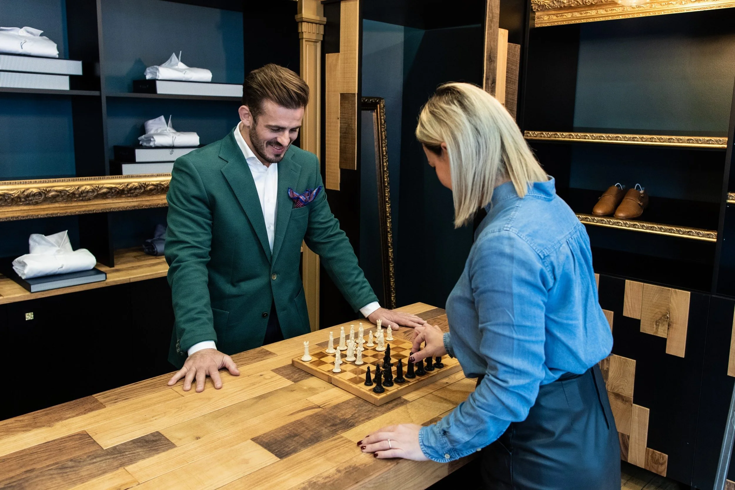 Deux personnes jouent aux échecs sur une table en bois dans une boutique ou un café, avec des étagères derrière elles contenant des boîtes et des chaussures.
