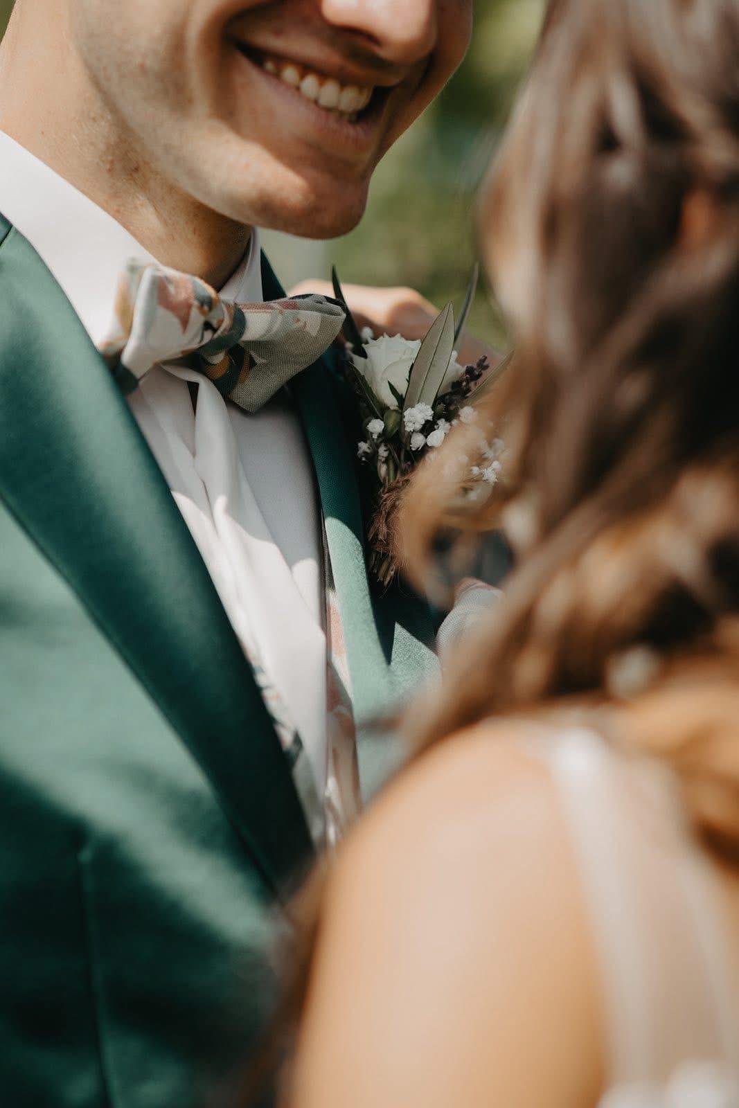 Un homme habillé en costume vert avec une veste et un nœud papillon floral, souriant lors d'un mariage, tenant une femme dont les cheveux bruns sont visibles, en arrière-plan il y a un bouquet de fleurs.