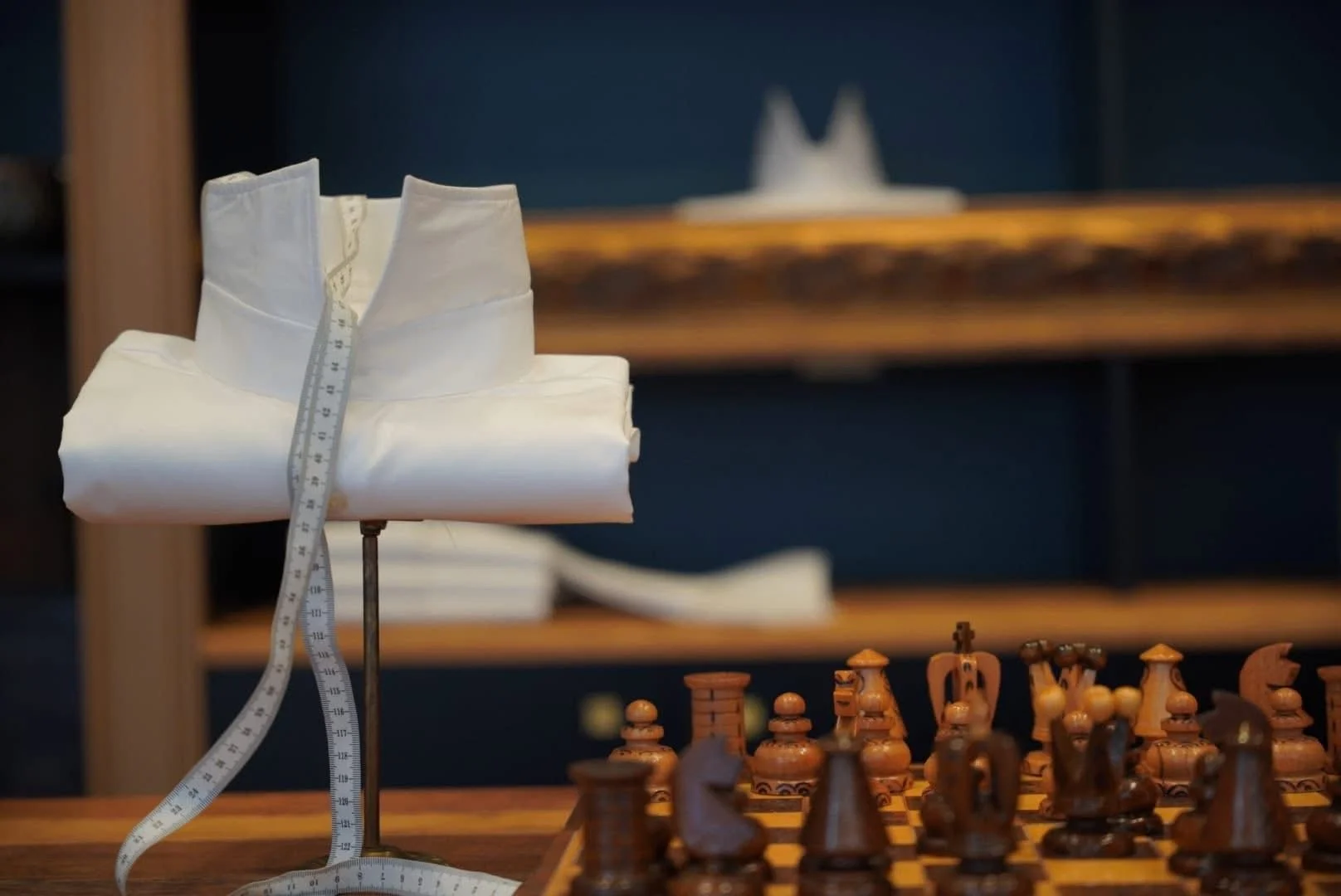 Une pièce de costume blanche avec un ruban à mesurer, placée sur une table de jeu d'échecs en bois avec des pièces en bois. En arrière-plan, une étagère en bois avec des objets blancs, dont une pièce de costume et un tissu blanc.