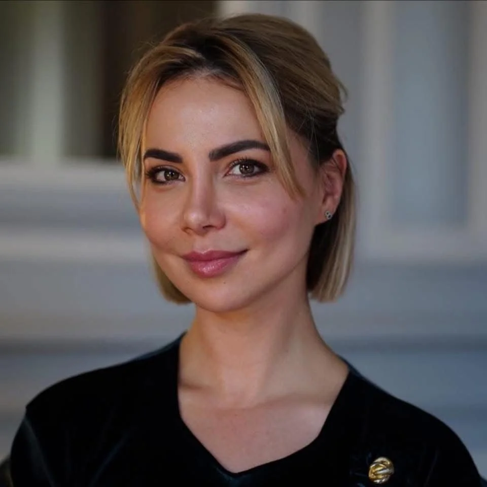 Portrait d'une femme avec des cheveux courts blonds, souriant, portant un haut noir, arrière-plan flou.