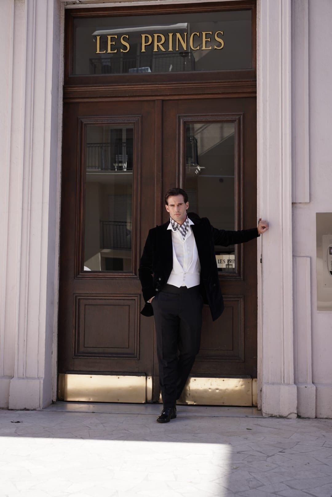 Un homme élégant en costume noir et blanc pose devant une porte en bois avec la mention "Les Princes" en lettres dorées.