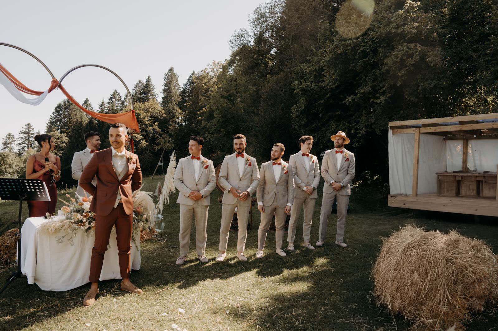 Groupe de personnes lors d'un mariage en extérieur, avec un homme en costume marron au premier plan, sept hommes en costumes beige et un homme en costume beige et un chapeau, tous portant une pochette rouge, dans un décor naturel avec herbe, arbres et décorations en paille et en tissus.