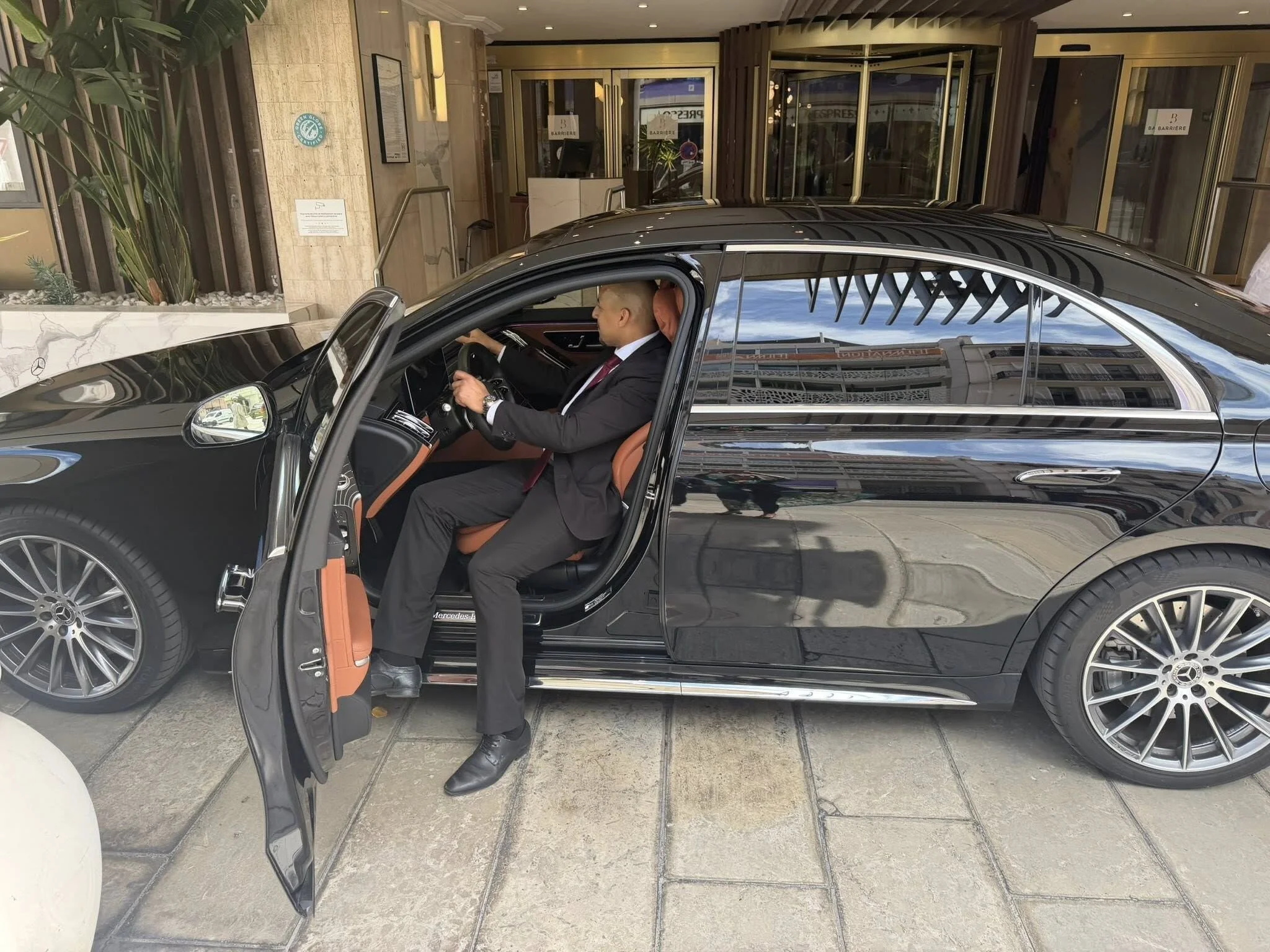 Homme en costume assis dans une voiture de luxe noire, entrée dans un hôtel ou un bâtiment avec un hall moderne, porte en verre et décor intérieur élégant.