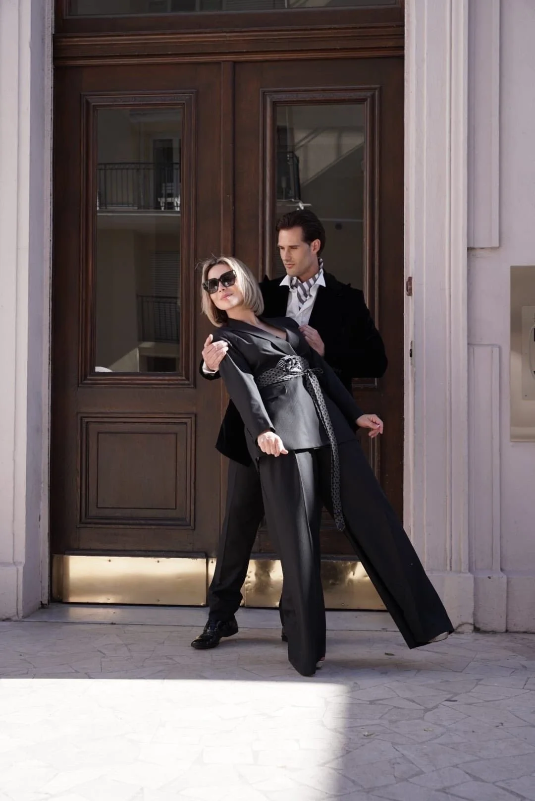 Un homme et une femme posant devant une grande porte en bois. La femme porte un costume noir avec une ceinture en dentelle et des lunettes de soleil, tandis que l'homme porte un costume noir avec une chemise blanche.