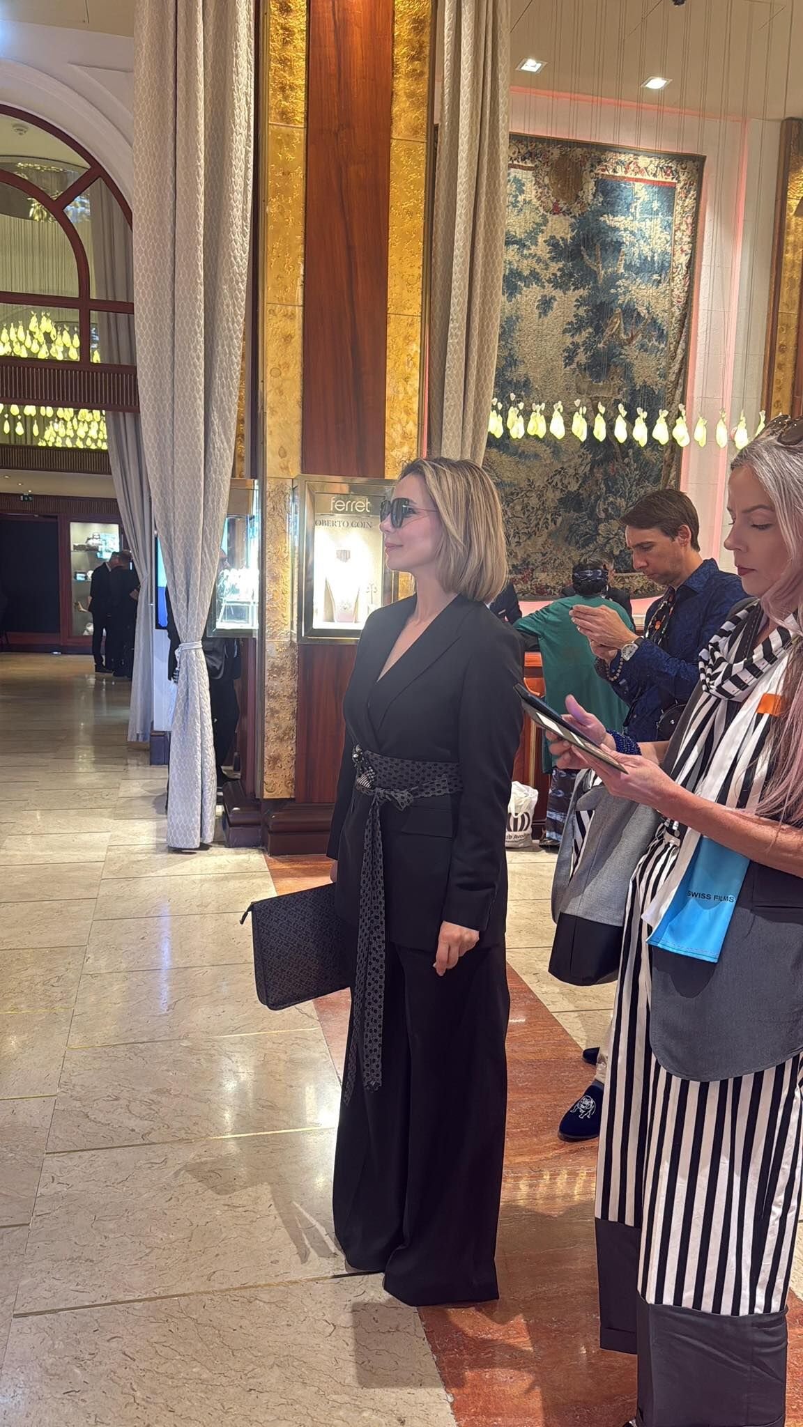 Une femme habillée en noir avec un chapeau noir, portant un sac noir et des lunettes de soleil, se tient dans un hall d'hôtel ou de centre d'affaires, entourée de plusieurs personnes.
