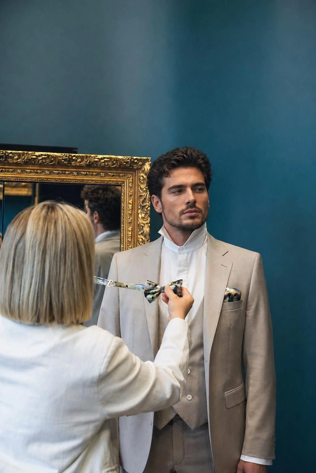 Une femme ajuste un nœud papillon à un jeune homme habillé d'un costume beige clair et d'une chemise blanche dans un miroir