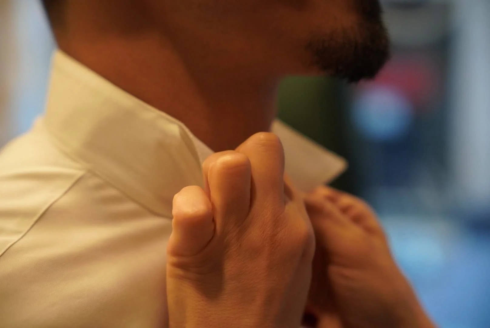 Un homme qui porte une chemise blanche ajuste ou soulève son épaule gauche avec ses mains. Le fond est flou et le focus est sur la main et le cou de l'homme.