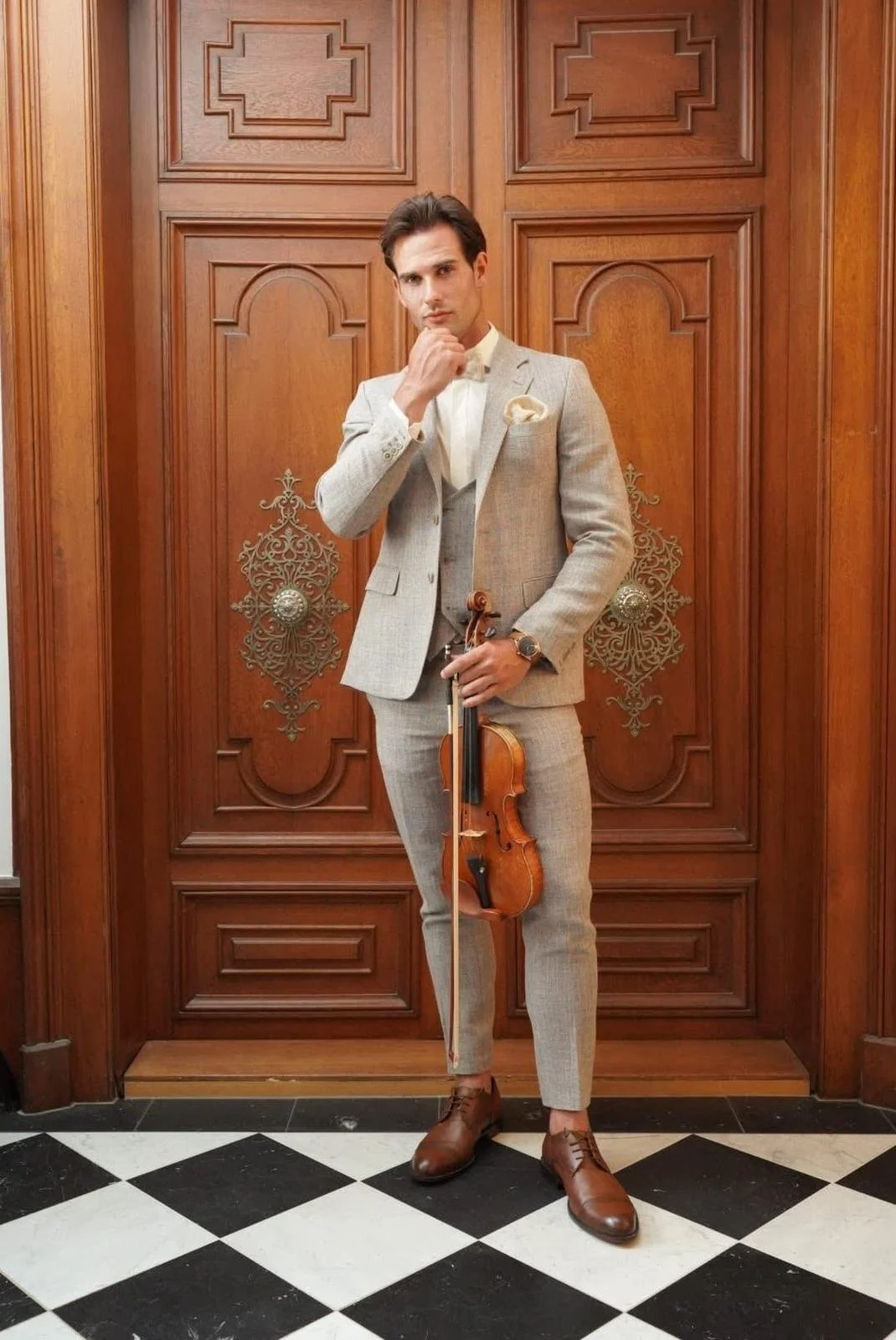 Un homme en costume gris, tenant un violon et un archet, se tient devant une porte en bois ornée de motifs en métal, dans une pièce élégante avec un sol en carreaux noir et blanc.