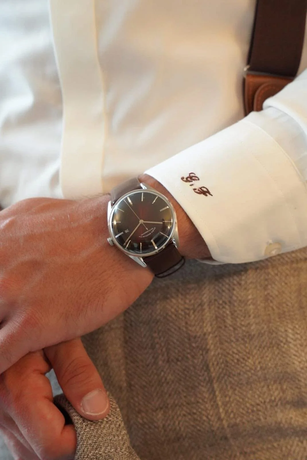 Un homme porte une montre élégante avec bracelet en cuir et chemise blanche. La montre a un cadran noir avec des aiguilles et des chiffres argentés.