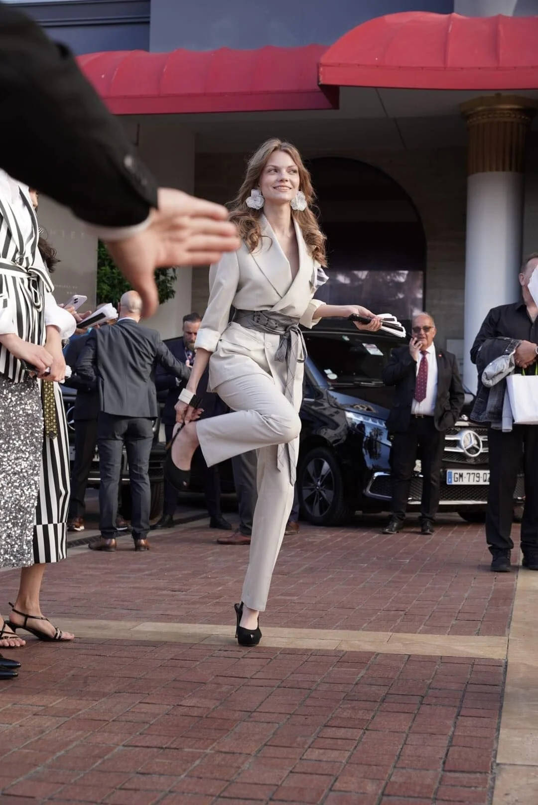 Une femme en costume beige, avec des boucles d'oreilles blanches, sourit en levant une jambe lors d'un événement public, entourée de plusieurs personnes, certaines portant des masques, devant des voitures de luxe. Elle semble marcher ou faire un gest