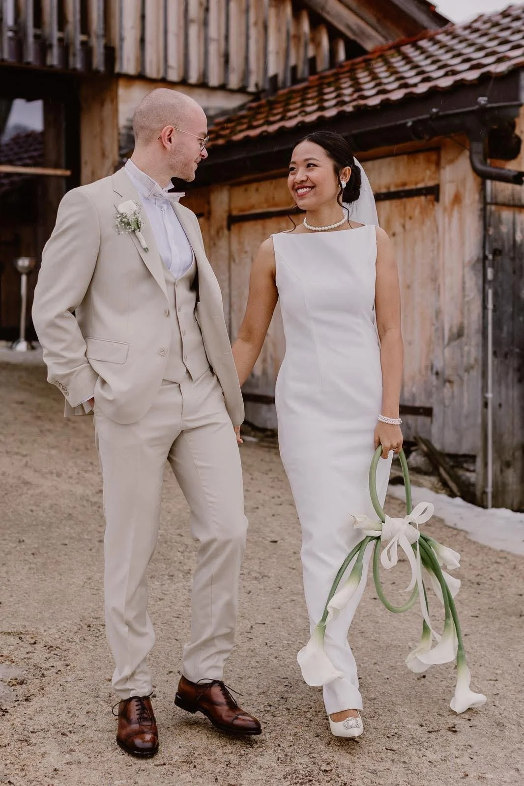 Un couple de mariés marche côte à côte dans une ferme en bois, la femme tient un bouquet de callas blanches et porte une robe blanche, l'homme porte un costume crème avec une boutonnière.