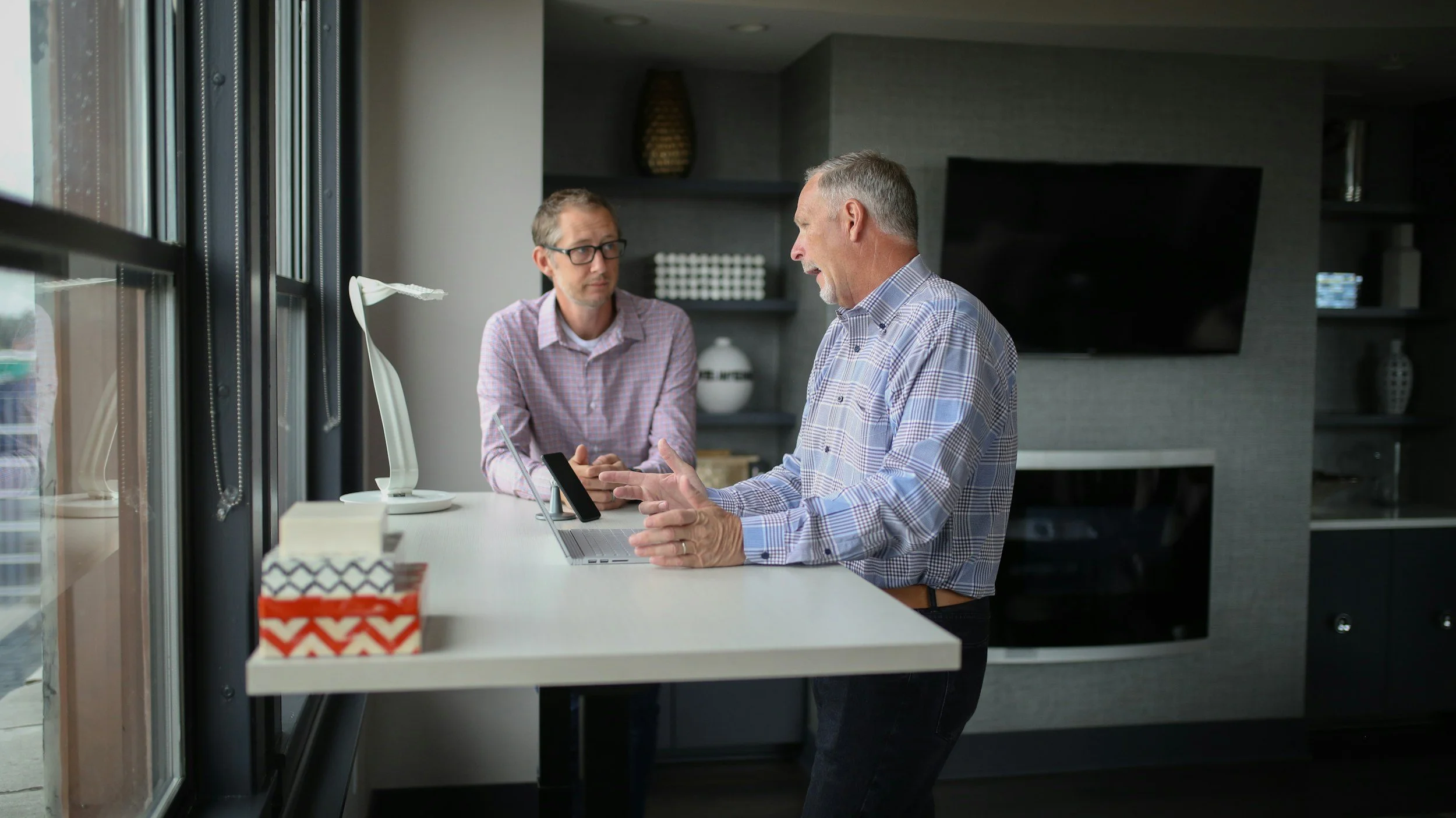 Two men having a conversation in a modern kitchen, with large windows, a laptop, and a smartphone on the counter.