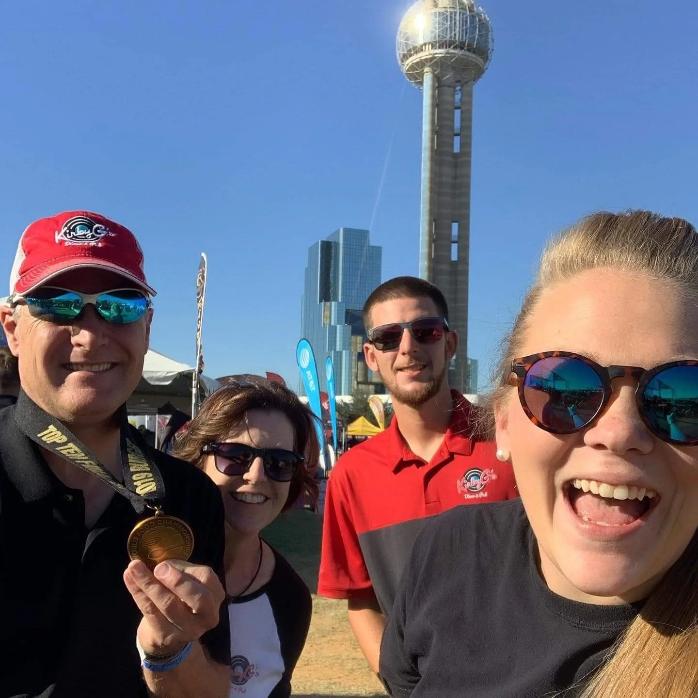Four people smiling for a selfie outdoors, with the AT&T Stadium in Arlington, Texas, in the background under a clear blue sky.