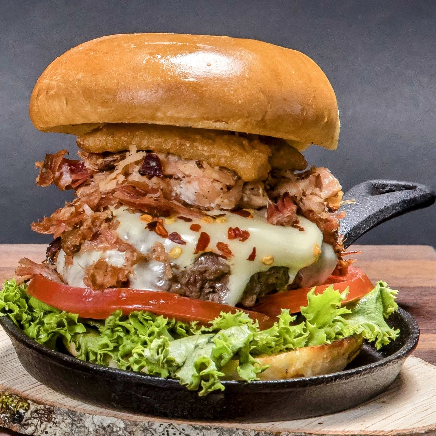 Close-up of a hamburger steak sandwich with cheese, onions, tomato, lettuce, and red pepper flakes served in a cast-iron skillet.