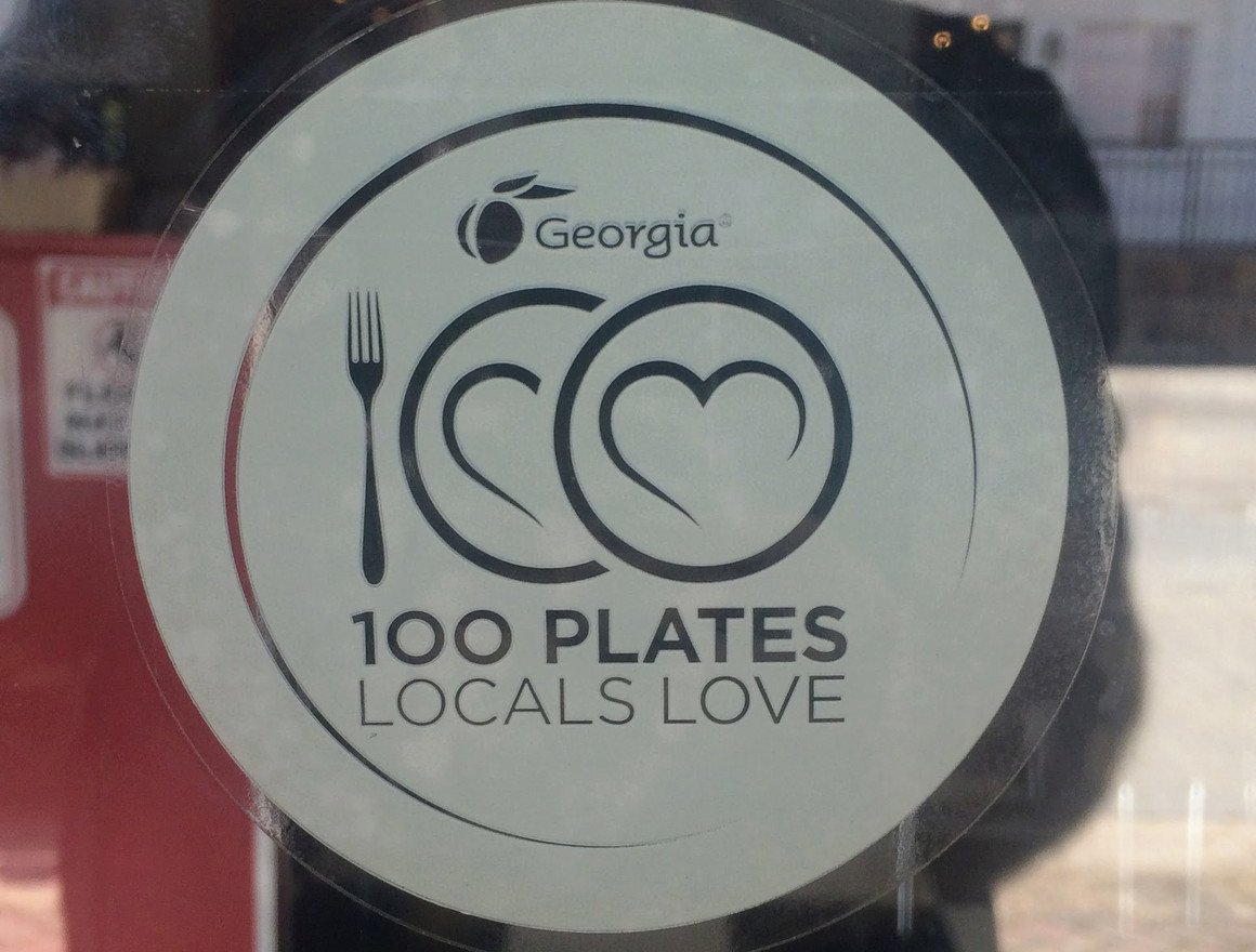 A sticker on a glass door or window with the Georgia state logo at the top, a fork icon next to a heart shape, and a circle with a heart shape inside it. The text reads "100 PLATES LOCALS LOVE."