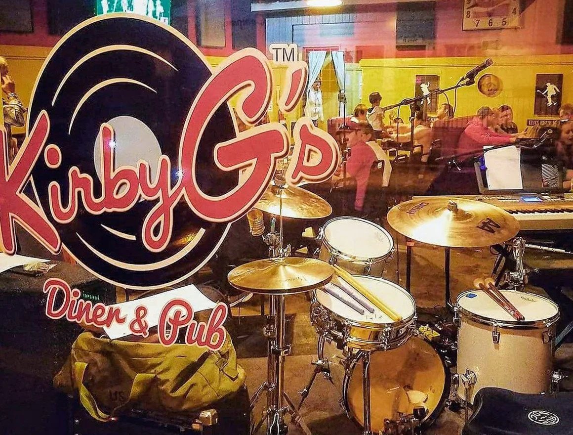 The window of a music venue or pub named Kirby G D, Diner & Pub, showing musical instruments including a drum set and a keyboard inside. The background shows patrons sitting at tables and socializing.