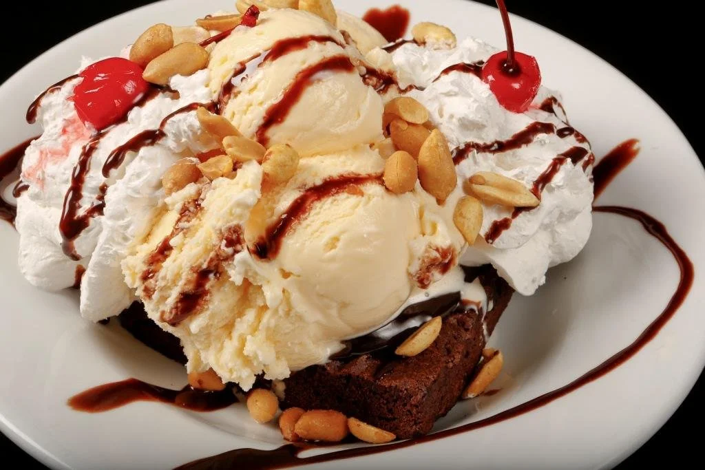 Ice cream sundae with vanilla ice cream, whipped cream, cherries, peanuts, chocolate syrup, and a brownie on a white plate.
