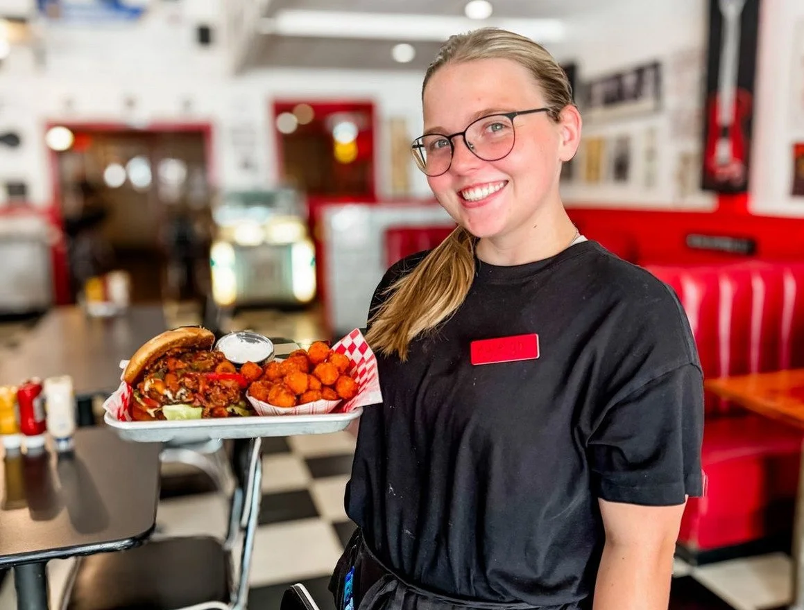 A smiling woman wearing glasses and a black shirt holding a tray with a burger, fried cheese balls, and a small container of sauce inside a retro-style diner.