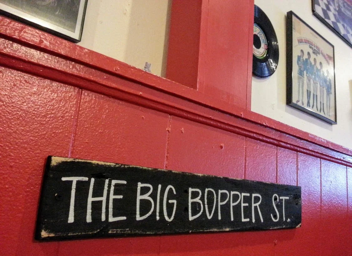 Close-up of a black wooden sign with white lettering that reads 'The Big Bopper St.' mounted on a red wall.