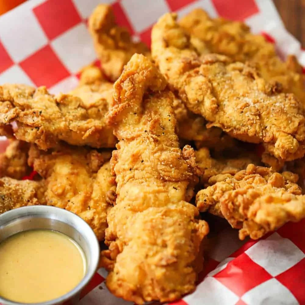 Fried chicken pieces with a side of honey mustard sauce on a red and white checkered paper basket liner.