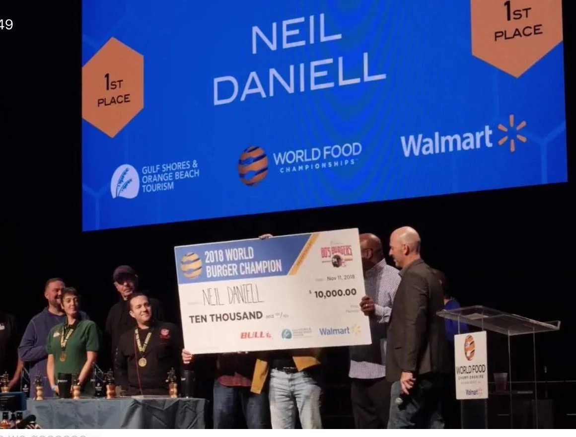 Neil Daniell holding a large check for $10,000 at the 2018 World Burger Championship awards ceremony, with judges and officials on stage, and a large screen behind displaying his name and first place victory.