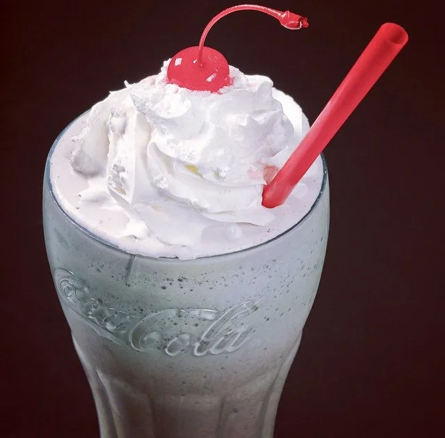 A milkshake in a Coca-Cola glass topped with whipped cream, a maraschino cherry, and a pink straw.