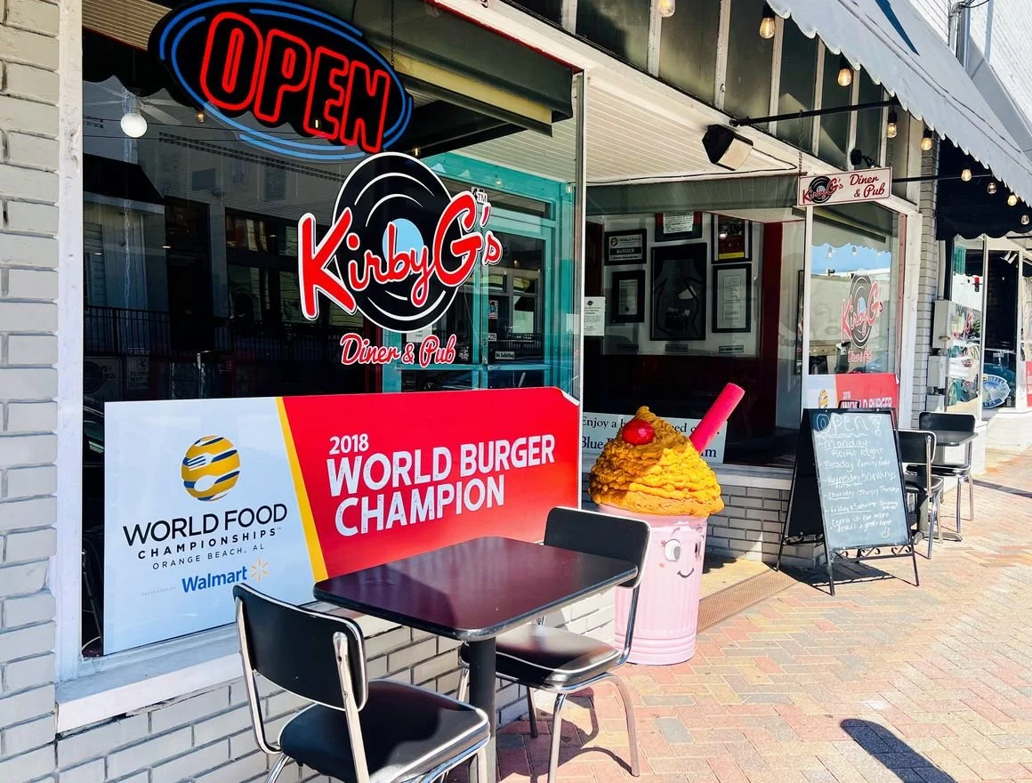 The storefront of Kirby G's Diner & Pub with an open sign, a large pink and yellow ice cream cone sculpture on the sidewalk, and signs advertising food and events outside.