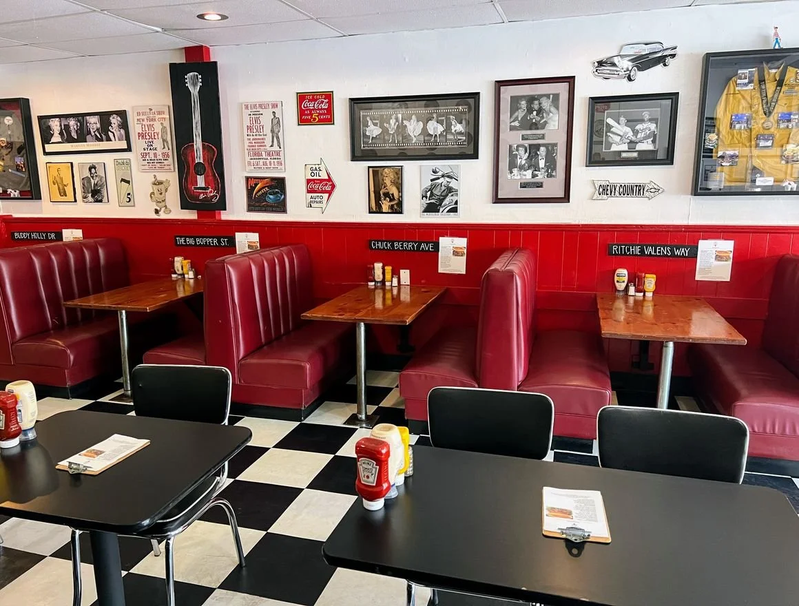 A retro-style diner with red leather booths, checkered black and white floor, and various framed pictures and posters on the white wall. There are condiment bottles on the tables and booths, and a wall display featuring a yellow racing jacket and a v