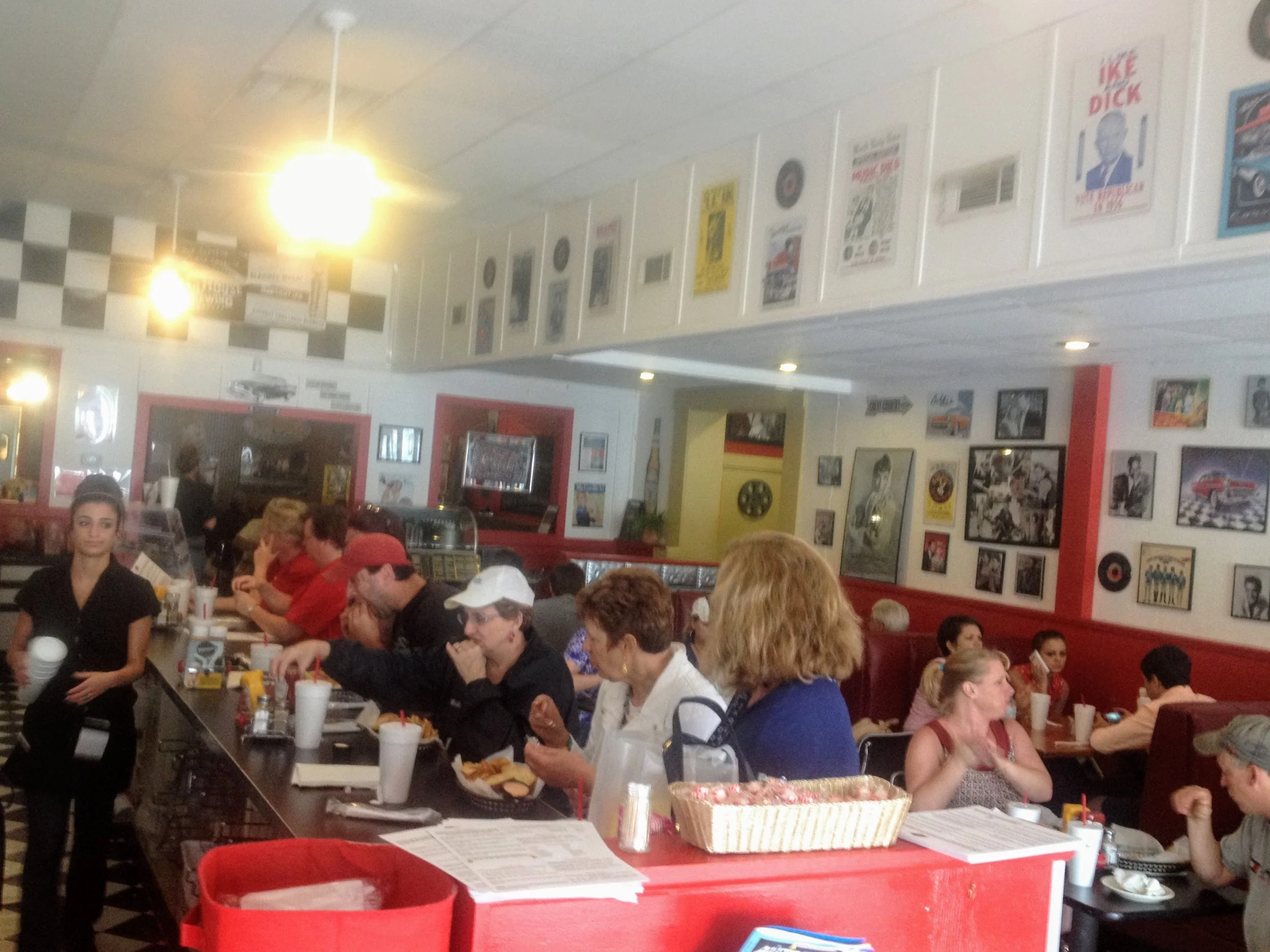 People sitting at a long table inside a diner or restaurant with vintage wall art and posters, some of them people, cars, and music related, with a woman in a black shirt standing in front.