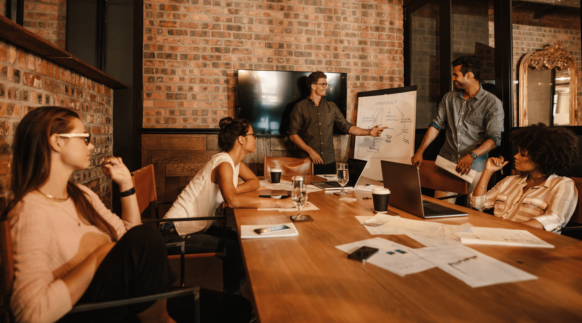 A group of five people are in a meeting room with wooden accents and exposed brick walls. Two men are standing near a whiteboard displaying strategy notes, while three women are seated at a table looking attentive. The table has laptops, papers, glasses of water, and coffee cups.