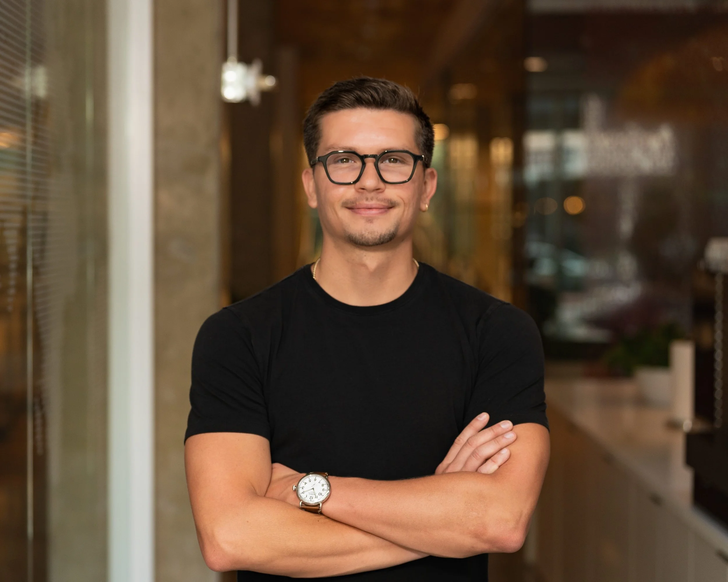 A young man with dark hair, glasses, and an earring is smiling at the camera with arms crossed, wearing a black t-shirt and a watch, standing in a well-lit indoor corridor.