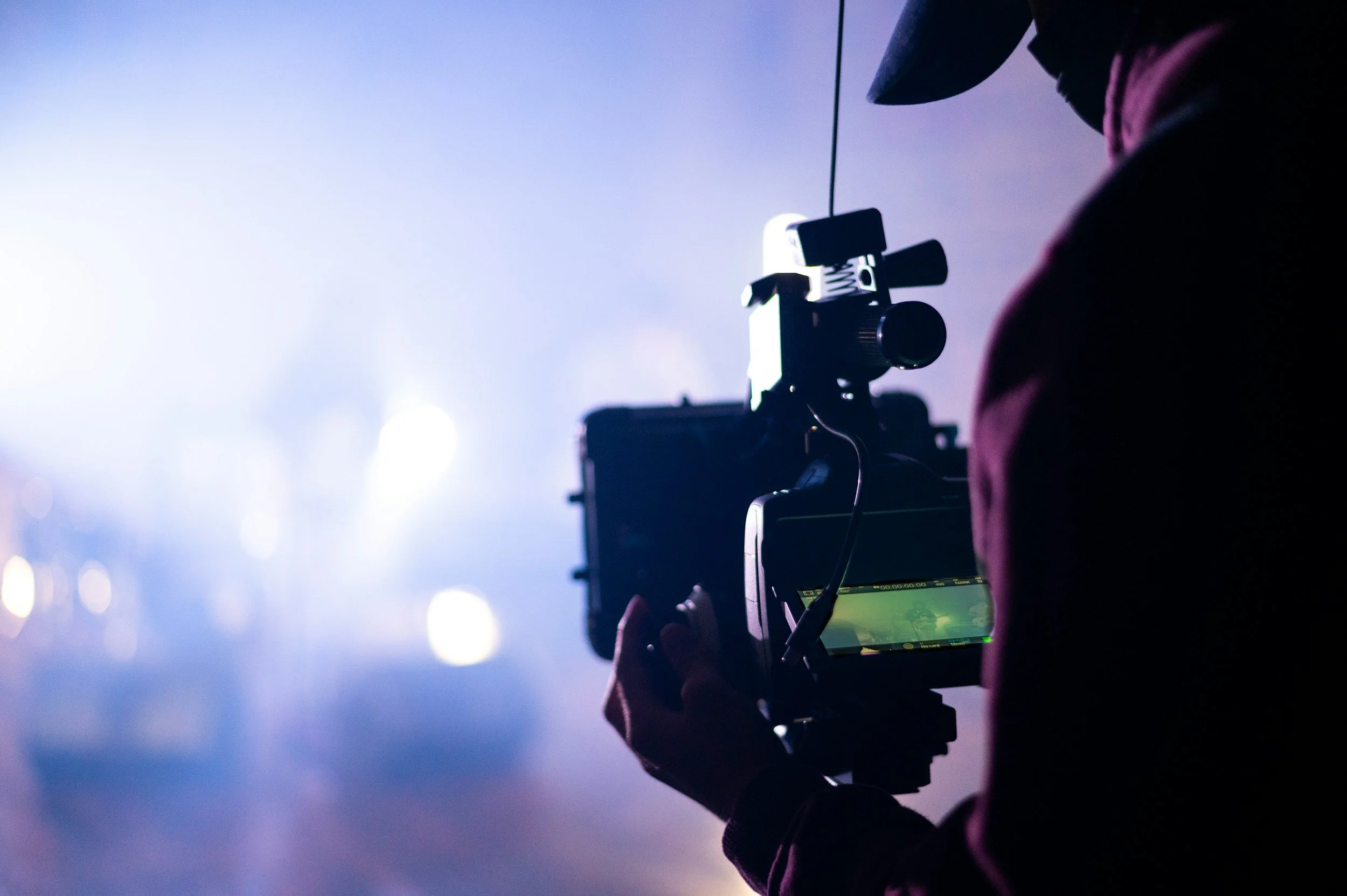 Person operating a professional video camera on a tripod in a low-light setting with blurred lights and a predominantly blue background.
