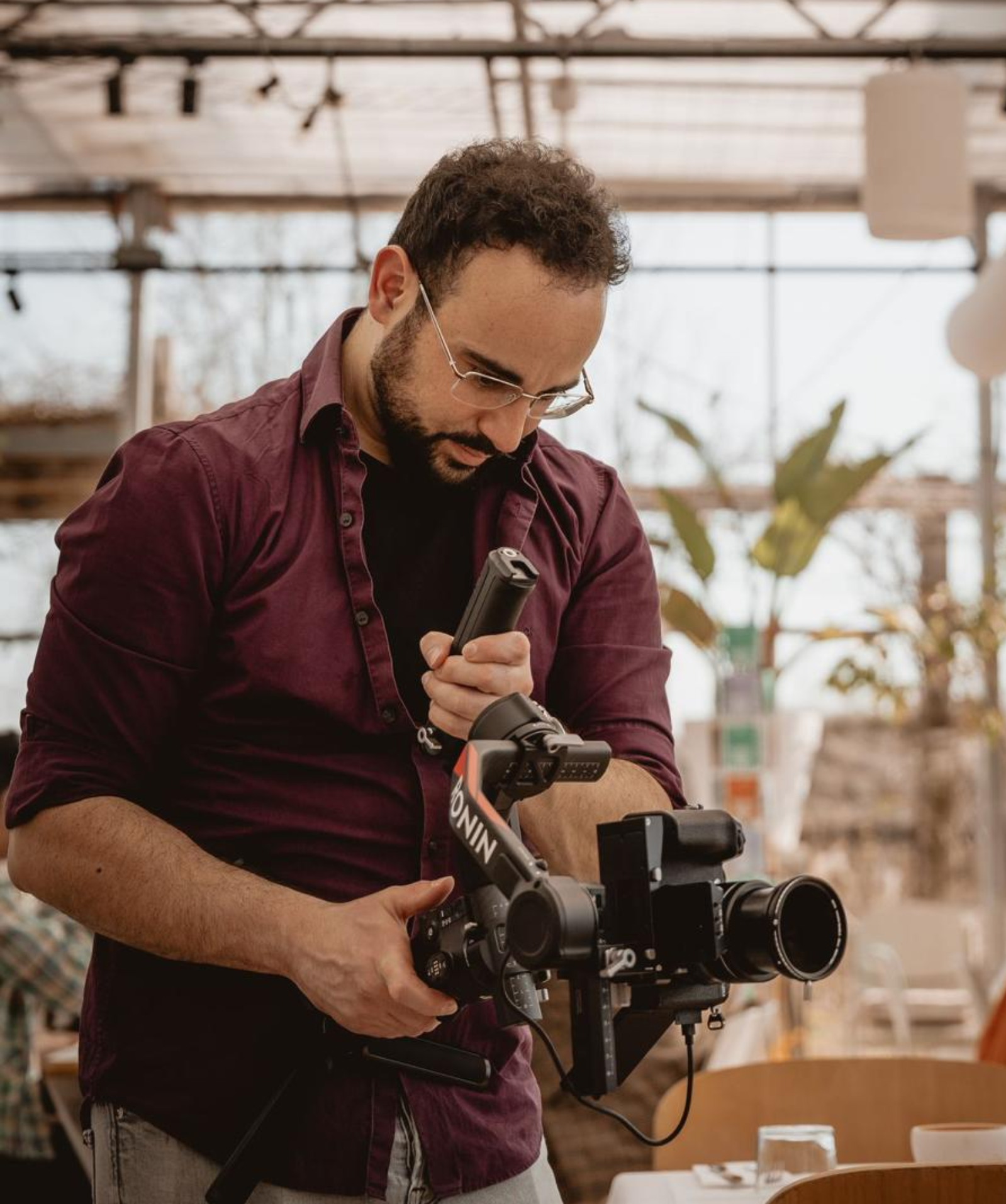 A man with curly hair and glasses operating a professional camera in a greenhouse or plant shop.