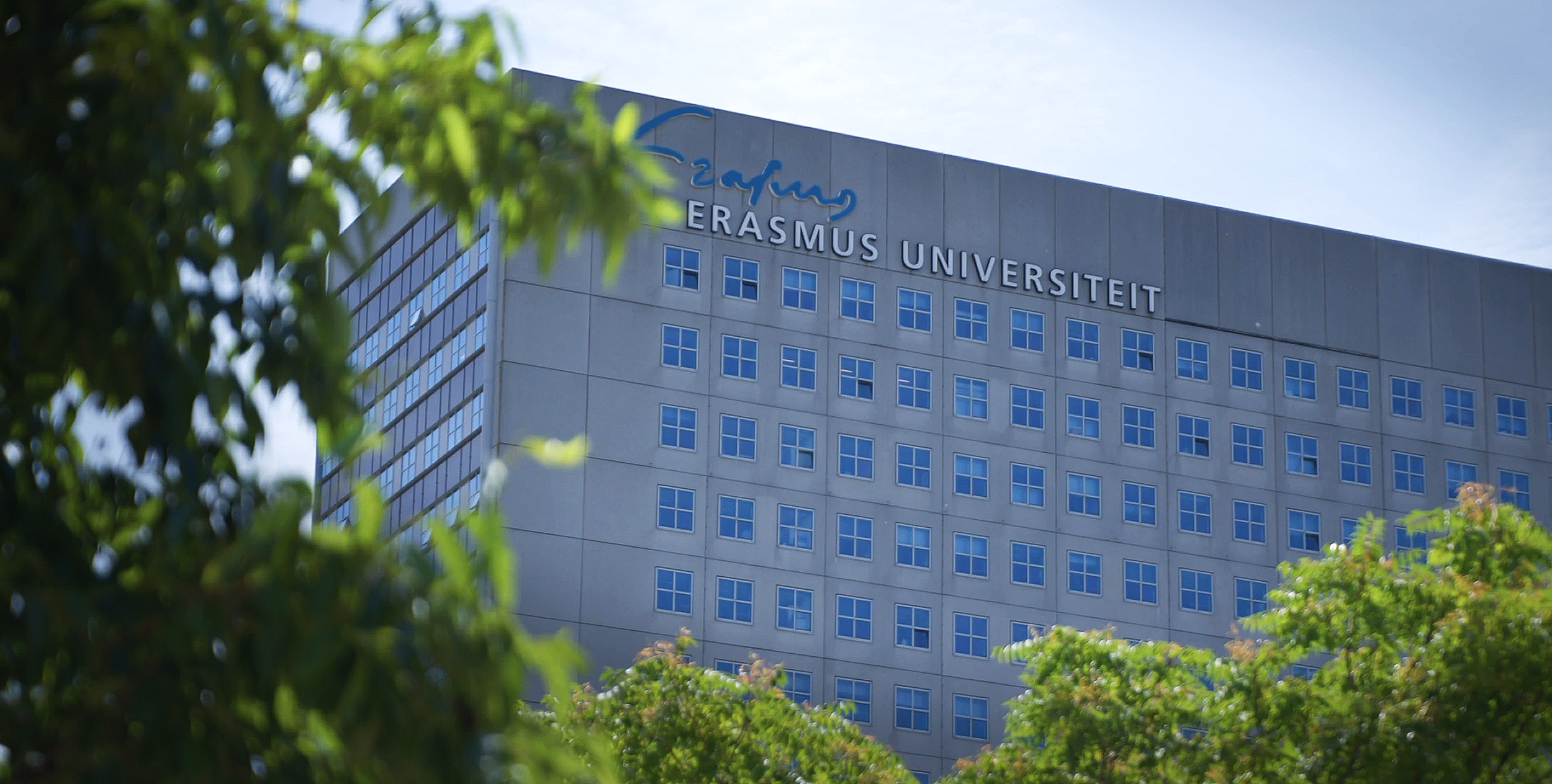 A tall building with blue windows and the words "Erasmus Universiteit" on the top, partially obscured by green trees in the foreground.